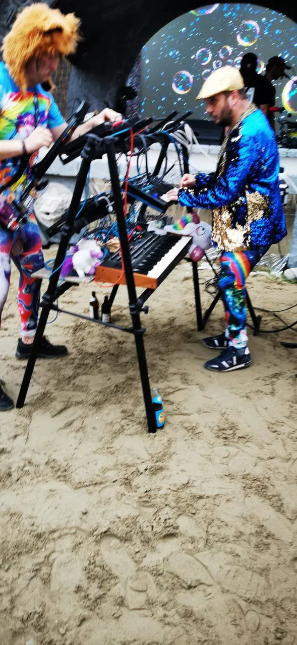 Glitter-clad performers with synths on a sandy festival floor